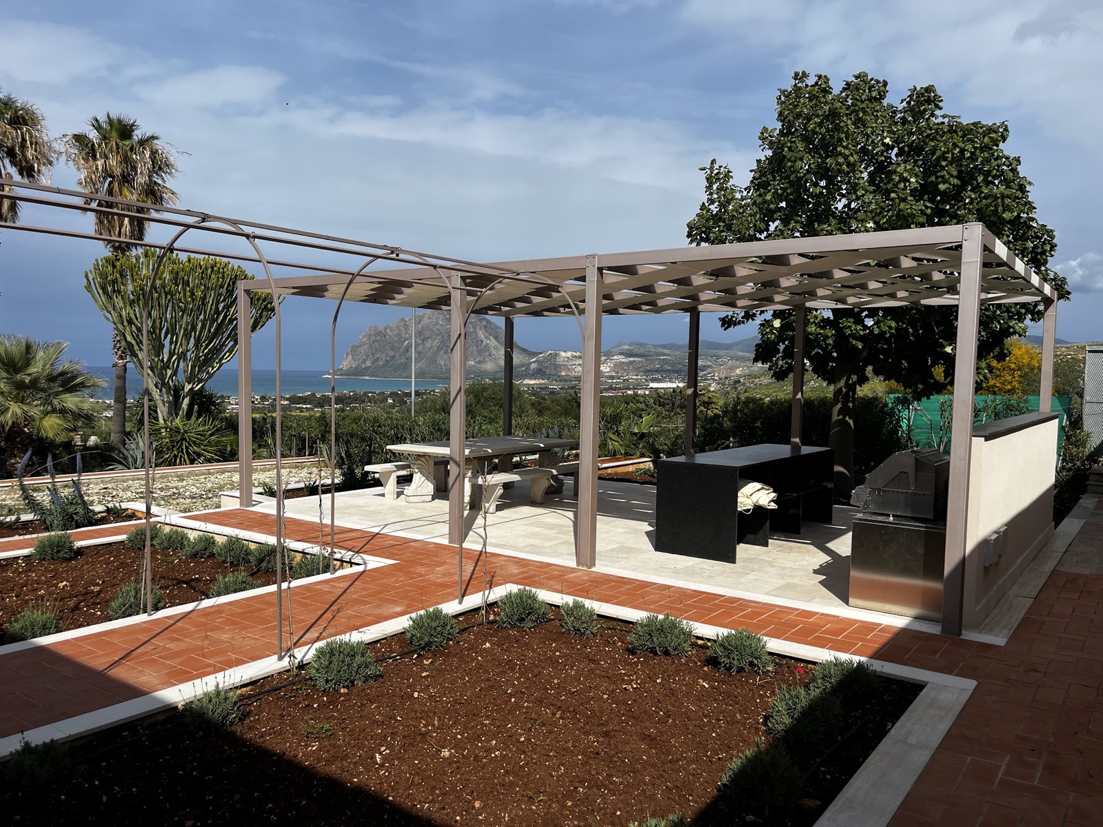 Pergola-shaded outdoor kitchen with built-in BBQ and stone dining table, Monte Cofano and the sea beyond